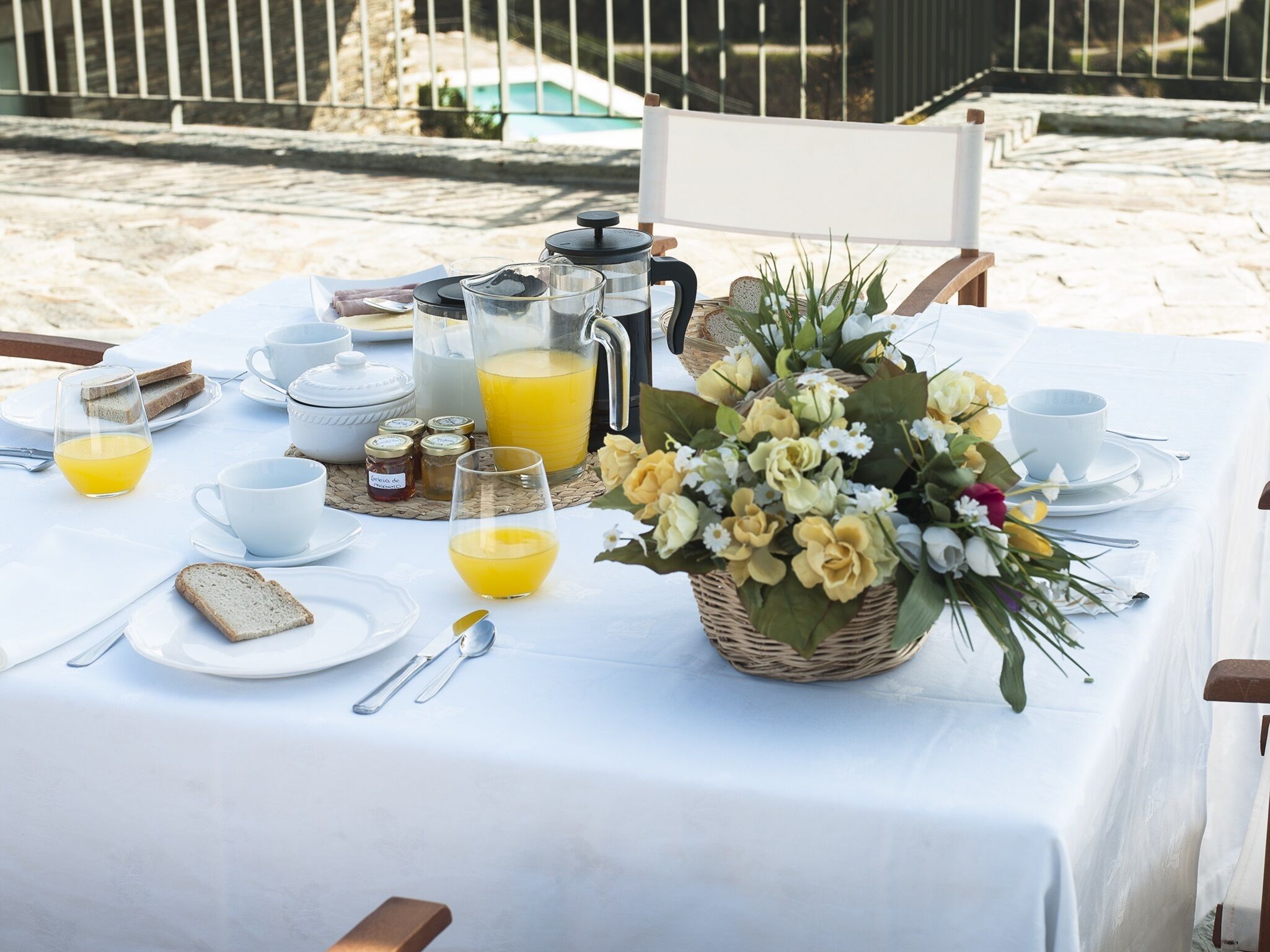 A breakfast setup on a beach table with a white tablecloth, including bread, butter, coffee, tea, and a flower arrangement.