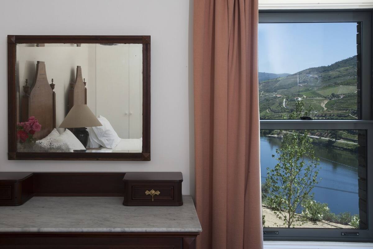 A room with a bed, a bedside table, a mirror, and a window with a view of a river and hills.