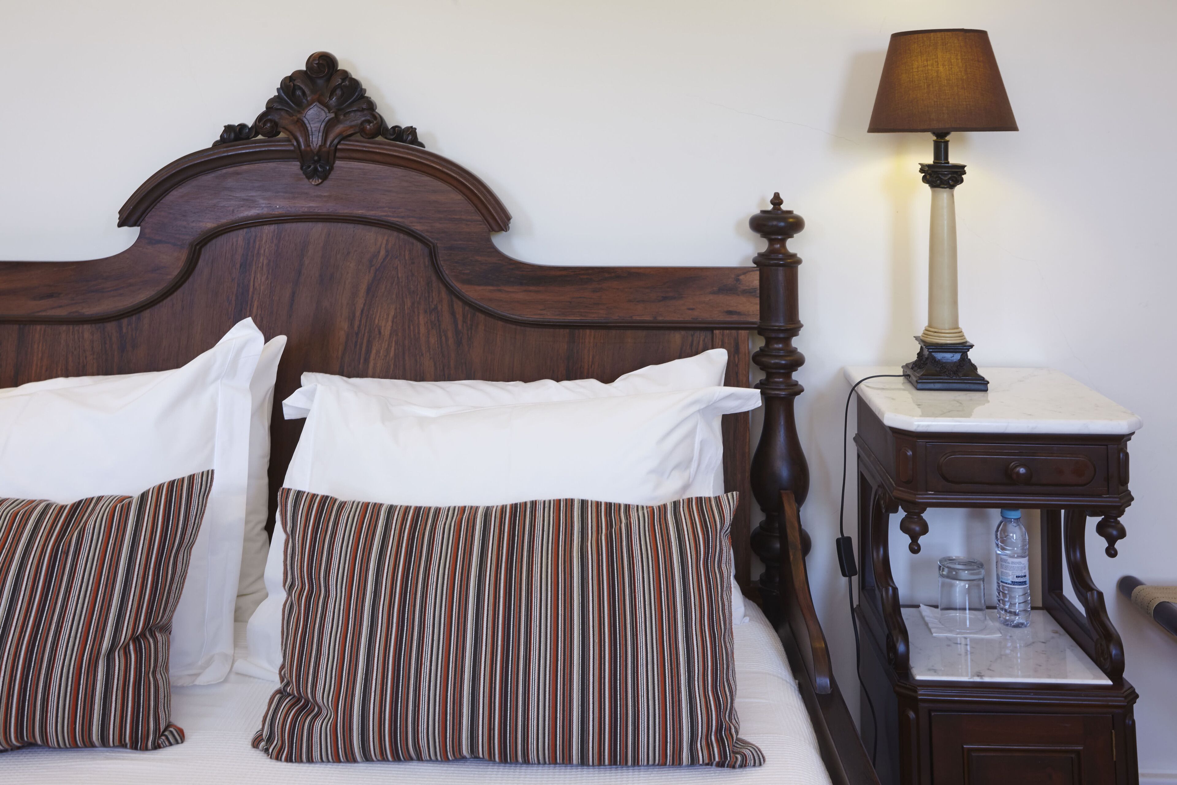 A bed with white linens and striped pillows, a wooden headboard, and a bedside table with a lamp and a bottle.