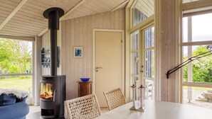 A cozy room with a wood-burning stove, a dining table, wicker chairs, and large windows offering a view of the outdoors. - 5 Person Holiday Home in Nakskov (Nakskov)