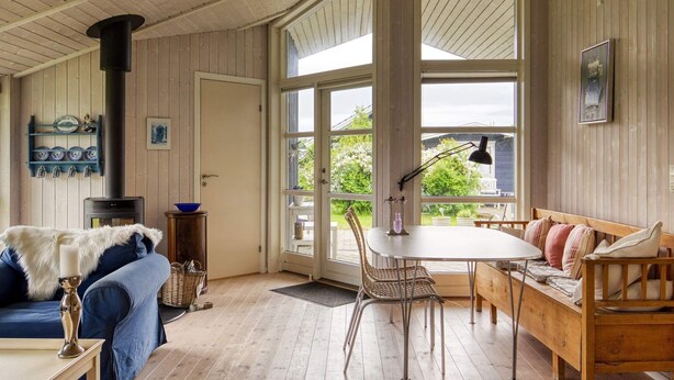 A cozy room with a wooden floor, a sofa, a table with chairs, a bench, a fireplace, and large windows. - 5 Person Holiday Home in Nakskov (Nakskov)