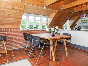 A dining area with a wooden table and chairs, a window with a view of greenery, and a kitchen area with modern appliances. - 6 Person Holiday Home in Ringkobing (Ringkobing)