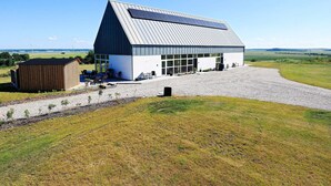 A modern building with a sloped roof and large windows, surrounded by a gravel driveway and grassy fields.