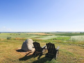 Two Adirondack chairs on a grassy field with a large rock and a view of a distant landscape.