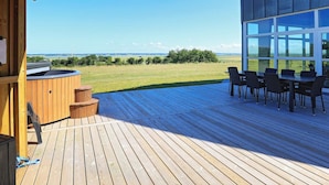 A wooden deck with a hot tub and outdoor seating area, overlooking a grassy field and wind turbines.