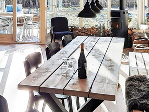 A wooden table with chairs, a bottle of wine, and glasses, with a large window and a wood stove in the background. - 5 Star Holiday Home in Dannemare (Dannemare)