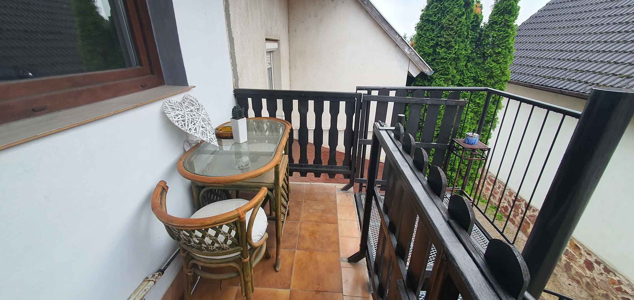 Ein kleiner Balkon mit einem Glastisch und zwei Korbstühlen, einem schwarzen Metallgeländer und Blick auf ein Haus und Bäume.