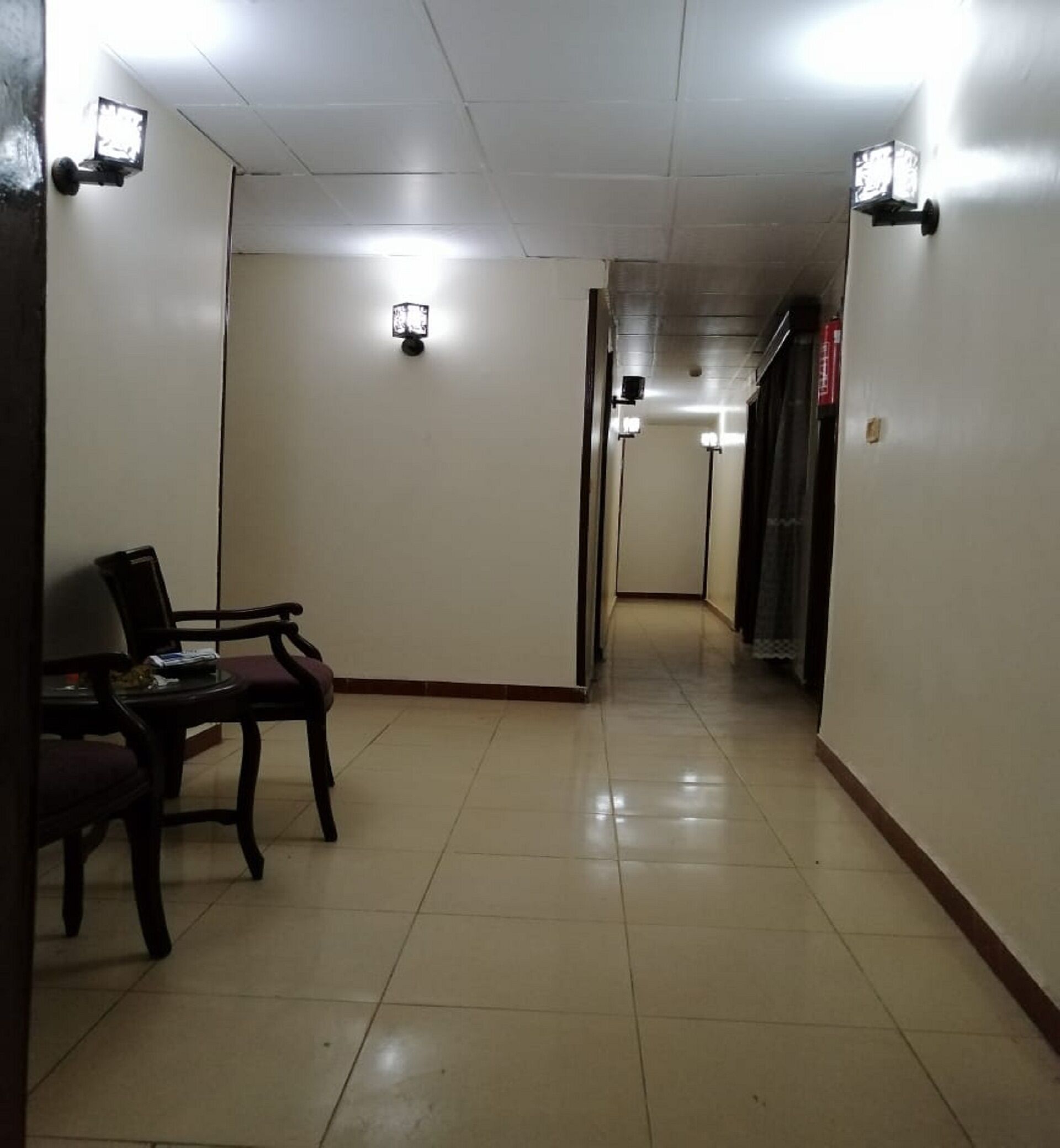 A hallway with a chair and a table, light fixtures on the walls, and a tiled floor.