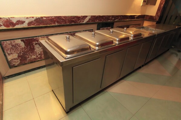 A buffet setup with four stainless steel chafing dishes on a counter, a marble backsplash, and a tiled floor.
