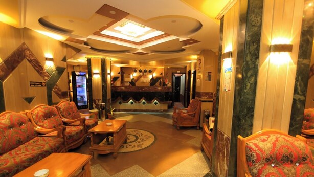 Een hotel lobby met een receptiebalie, een zithoek met sofa’s en fauteuils, een salontafel en een decoratief plafond.