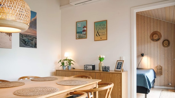 A dining area with a wooden table and chairs, a hanging light fixture, and wall-mounted artwork. - 4 Star Holiday Home in Nexo (Nexø)