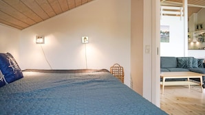 A bedroom with a bed, a bedside lamp, a wall-mounted light, and a view of a living area with a sofa and a table. - 4 Star Holiday Home in Nexo (Nexø)