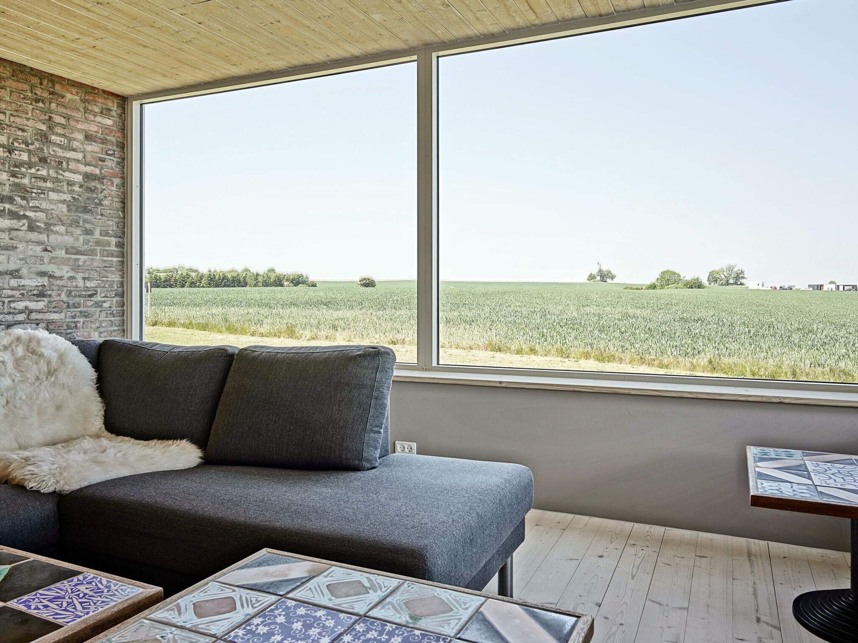 Una habitación con un sofá gris, un suave plaid blanco y vistas a un campo verde a través de amplios ventanales.