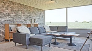 A modern living room with a sectional sofa, a wooden floor, a brick wall, and a large window offering a view of a field.