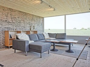 Un salon moderne avec un canapé d’angle, un plancher en bois, un mur en briques et une grande fenêtre donnant sur un champ.