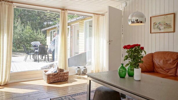 A room with a large sliding glass door leading to an outdoor patio, a sofa, a table with a vase of flowers, and a hanging light fixture. - 6 Person Holiday Home in Aakirkeby (Akirkeby)