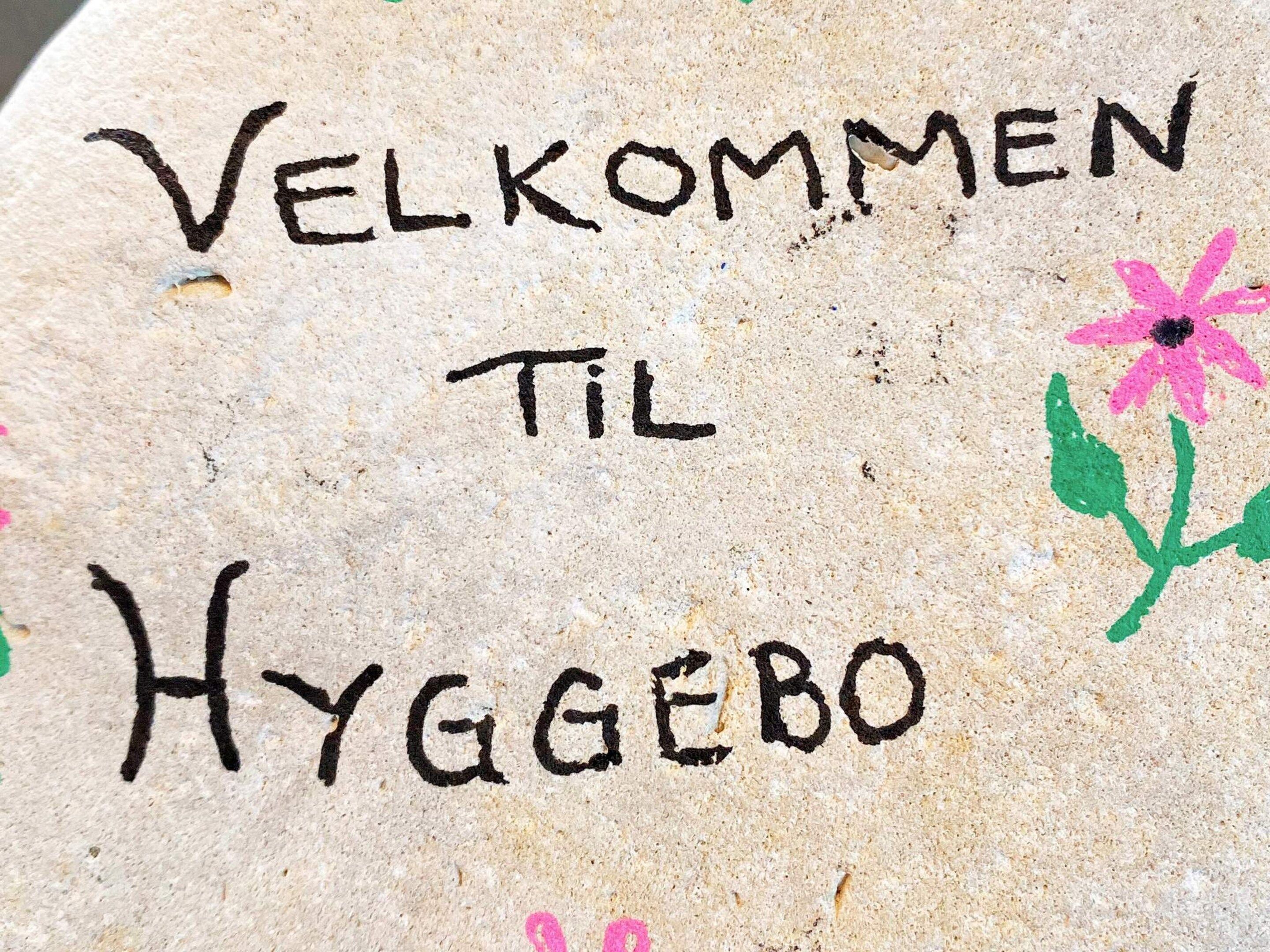 A stone with 'VELKOMMEN TIL HYGGEBO' written on it, accompanied by a pink flower drawing.