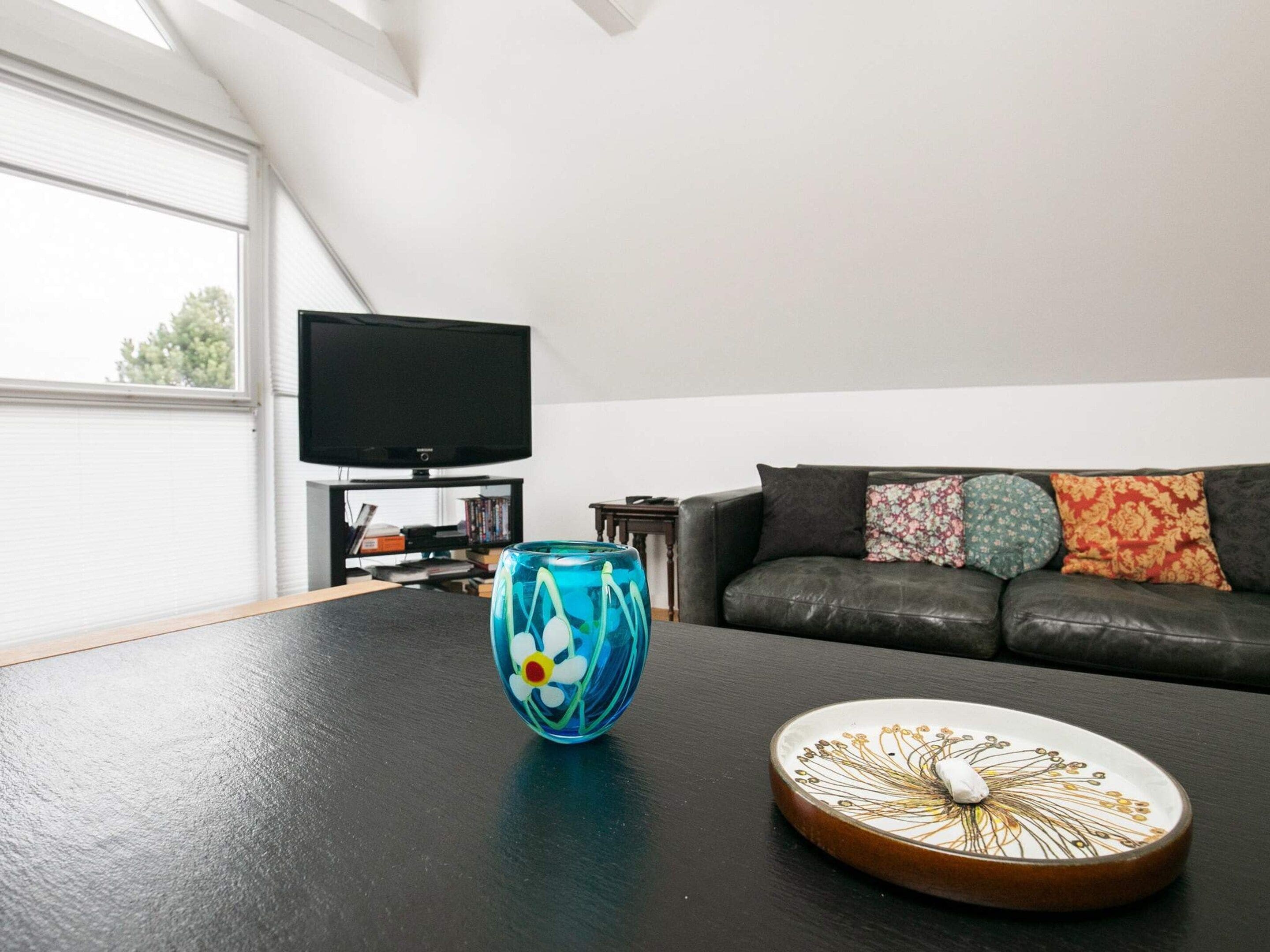 Un salon avec un canapé en cuir noir, un téléviseur posé sur un meuble et un vase en verre décoratif sur une table.