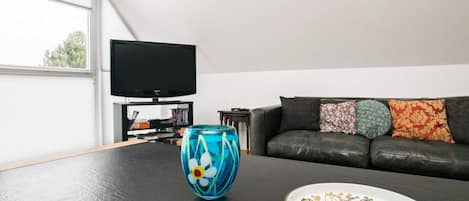 Una sala de estar con un sofá de cuero negro, un televisor sobre un mueble bajo y un jarrón de cristal decorativo sobre una mesa.