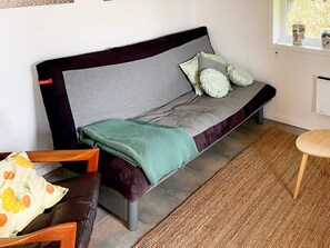 A single bed with a grey headboard, a green throw blanket, and two pillows. There is a small wooden table with a chair to the left and a small round table to the right.