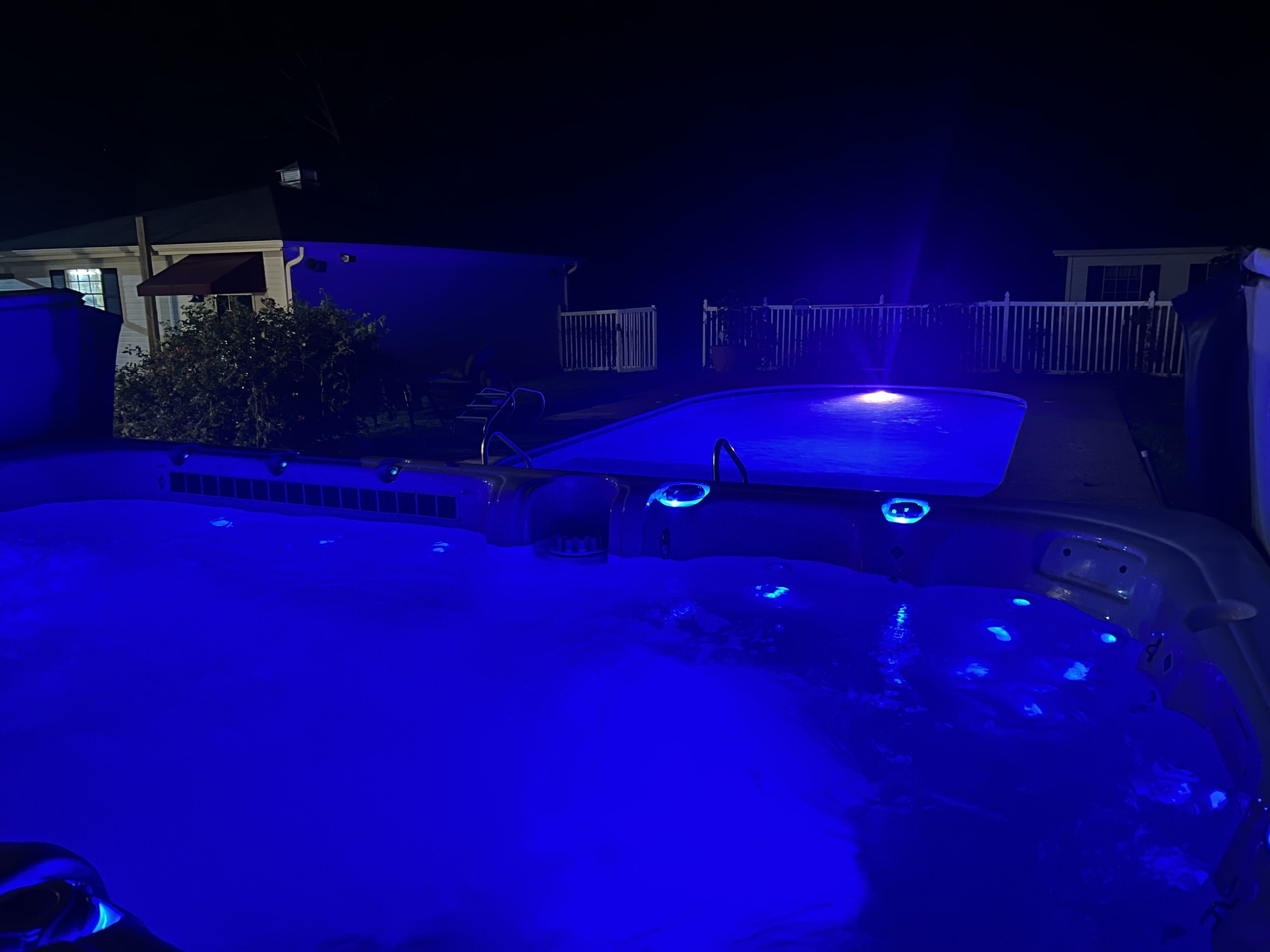 A hot tub with blue lighting, surrounded by a white fence and a house in the background.