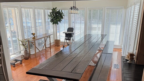 A spacious dining area with a long wooden table and benches, large windows with blinds, a ceiling fan, and a potted plant.