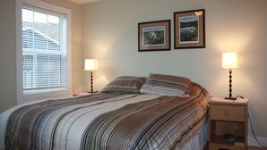 Une chambre avec un grand lit, deux tables de chevet avec des lampes, une fenêtre avec des stores et deux tableaux encadrés au mur.