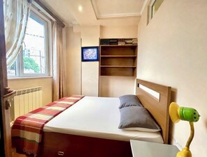 A bedroom with a bed, a shelf, a window, and a wall-mounted TV.