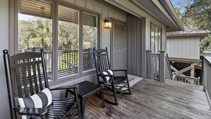 A covered porch with two rocking chairs and a table, large windows with blinds, and a wooden deck.