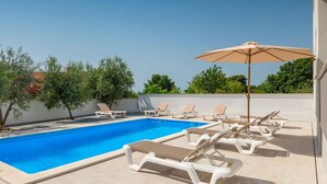 A swimming pool with lounge chairs and an umbrella, surrounded by greenery and a clear sky.