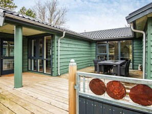 Una terraza de madera con una mesa, sillas, un ventanal de vidrio y una barandilla de metal con troncos de madera.