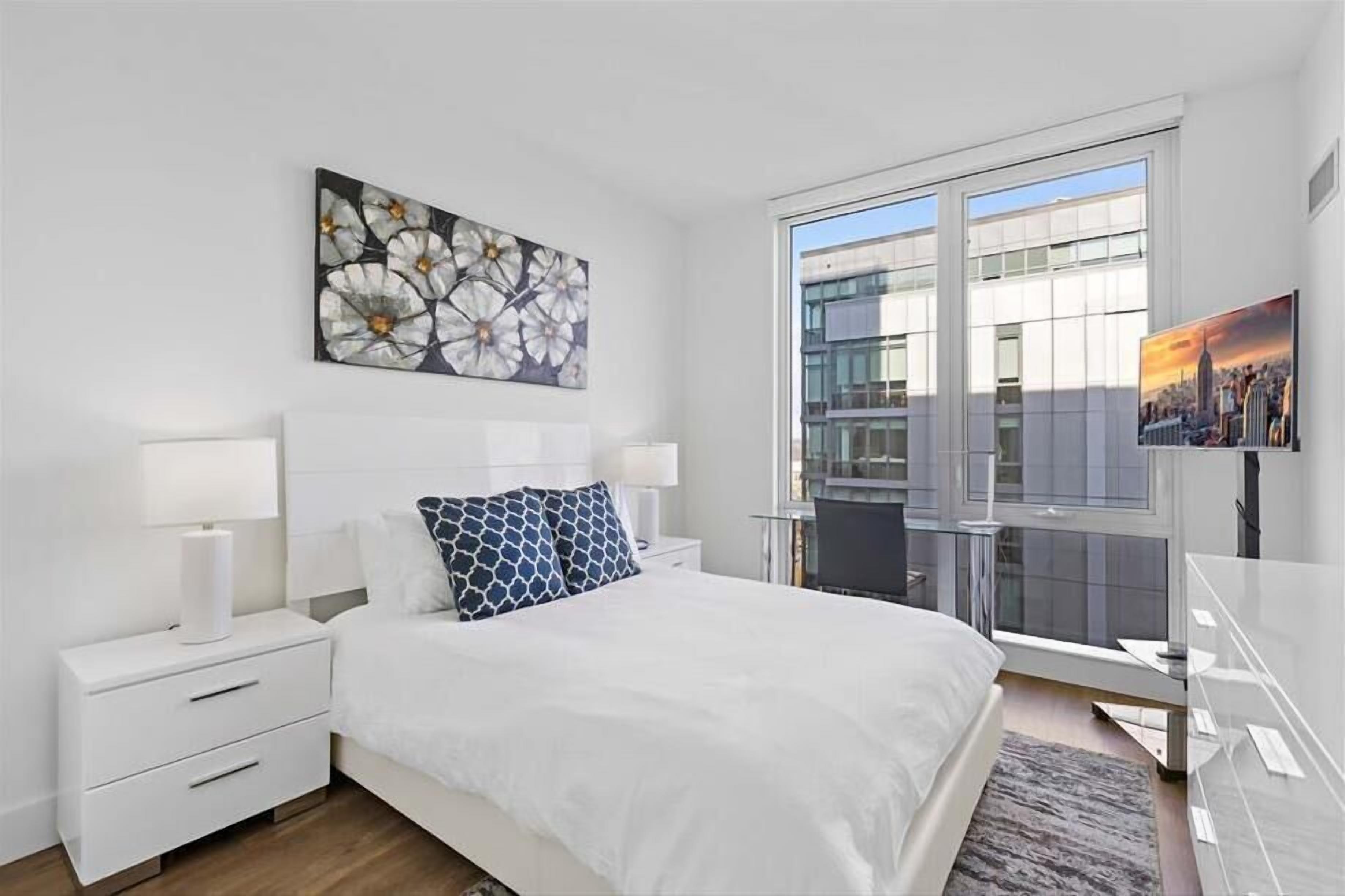 Un dormitorio moderno con una cama grande, mesitas de noche blancas, un cuadro de un paisaje urbano y vistas de un edificio a través de una puerta de vidrio.