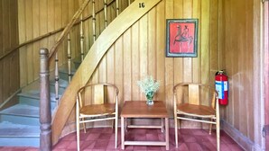 Un escalier en bois portant le numéro 16, deux chaises en bois, une petite table avec un vase et un extincteur fixé au mur.
