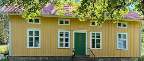 A yellow building with a green door and windows, surrounded by grass and trees.