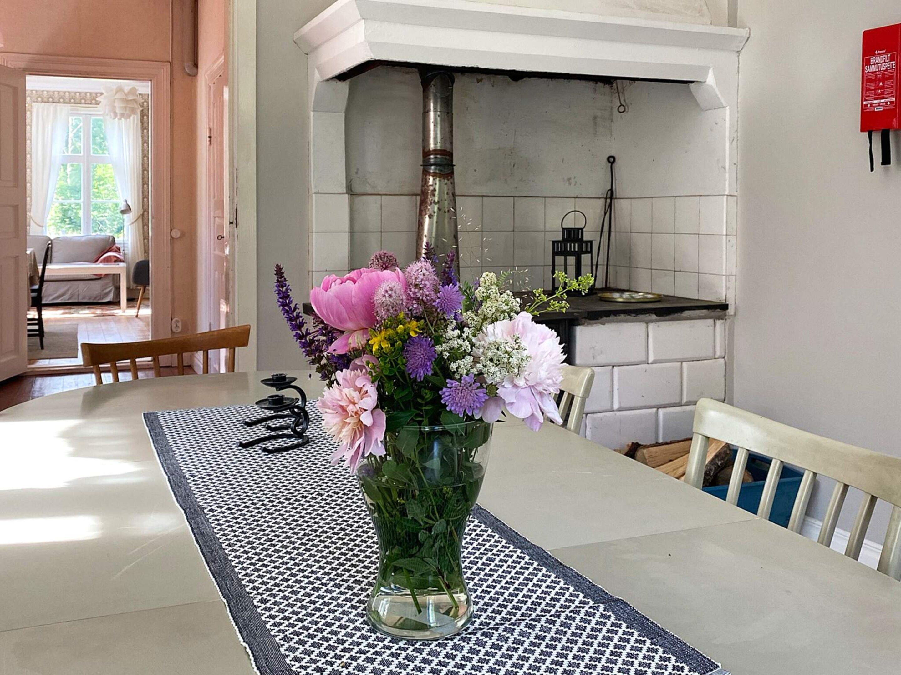 Un comedor con una mesa lista para dos, un jarrón de flores y una cocina al fondo.