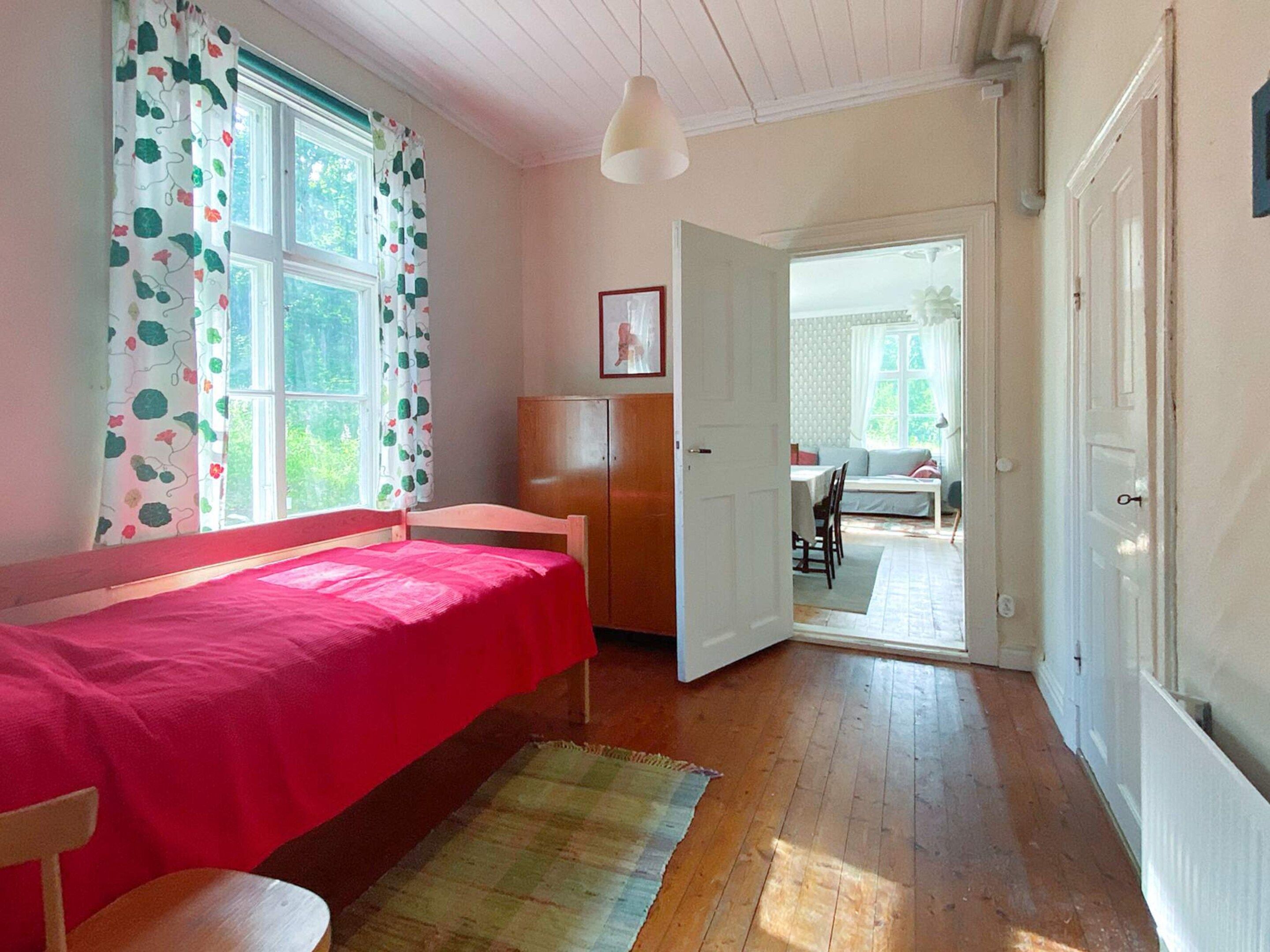 A room with a bed, a wooden wardrobe, a door leading to another room, and a window with curtains.