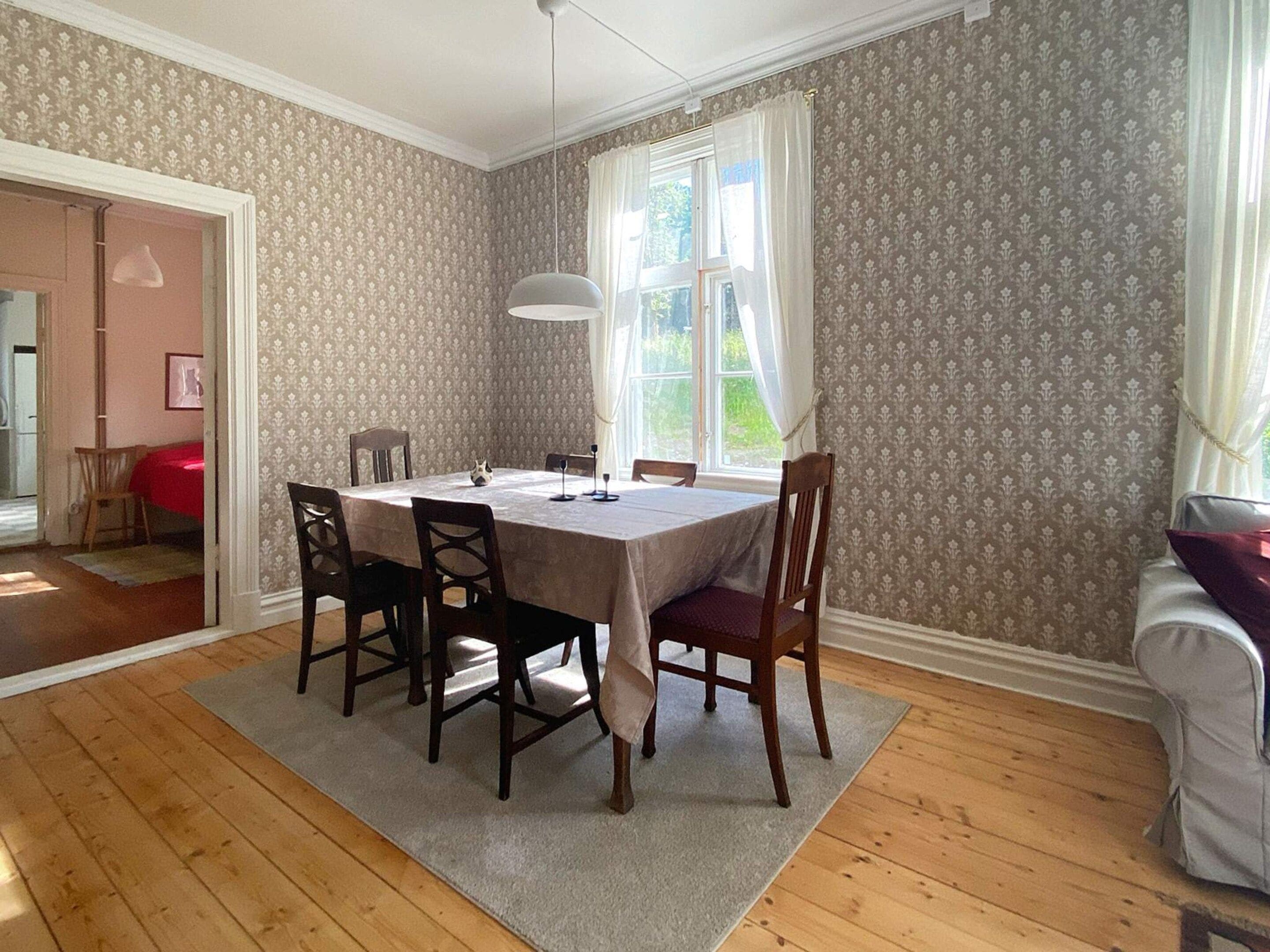 A dining room with a table set for four, surrounded by chairs, a chandelier, and a window with curtains.