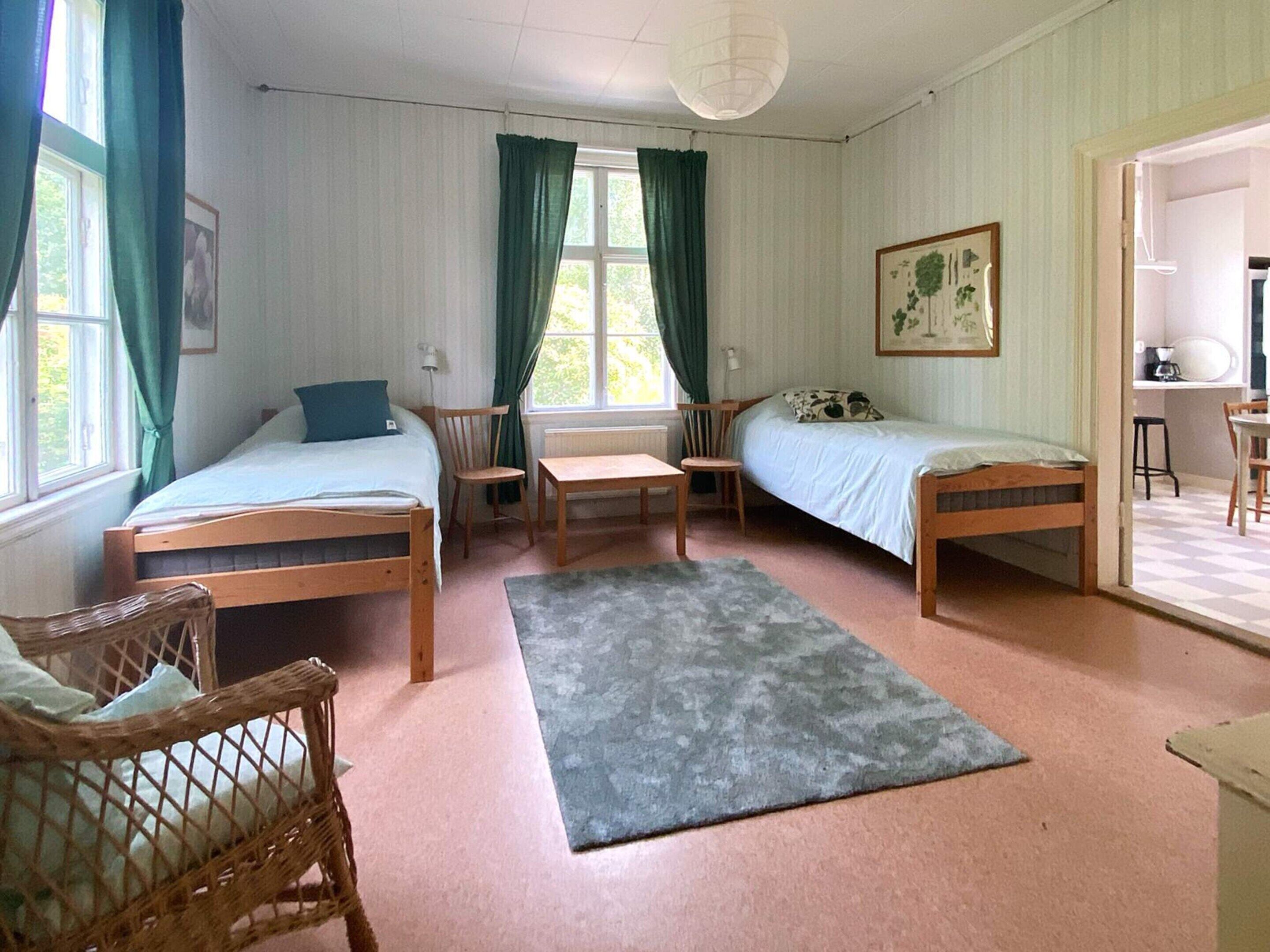 A room with two single beds, a wooden chair, a small table, and a window with curtains.