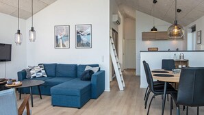 Un salon moderne avec un canapé d’angle bleu, un coin repas avec une table et des chaises en bois, et une cuisine dotée de luminaires suspendus.