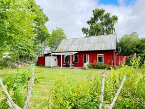 A red barn with a covered porch, surrounded by lush greenery and a wooden fence. - 4 Person Holiday Home in Hogklint (Visby)