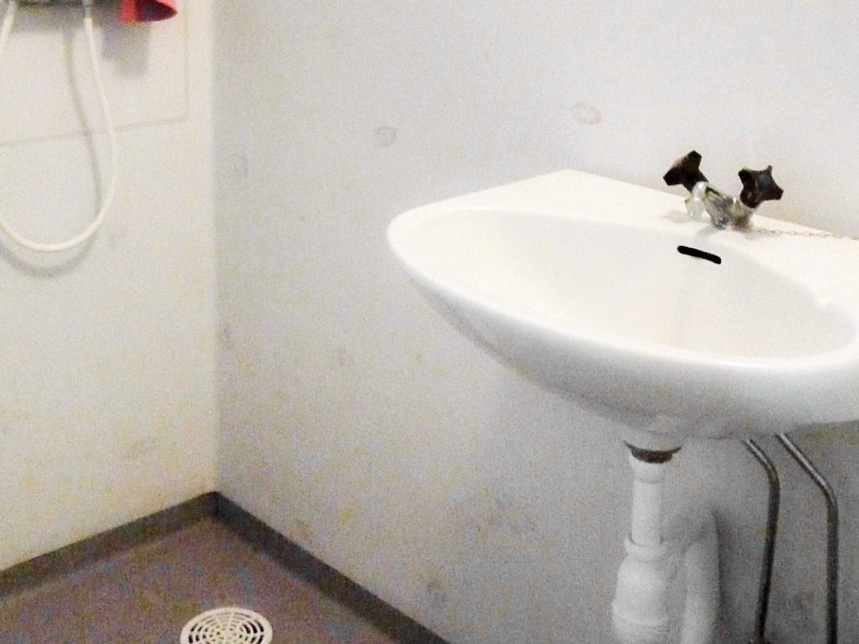 Un lavabo blanc, avec un seul robinet, placé devant un mur blanc et au-dessus d’une grille d’évacuation au sol.