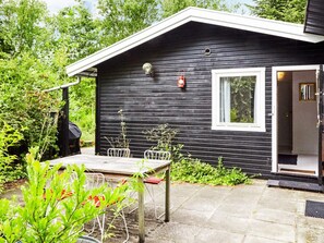 Een zwarte houten blokhut met één raam, een deur en een klein terras met een tafel en stoelen.