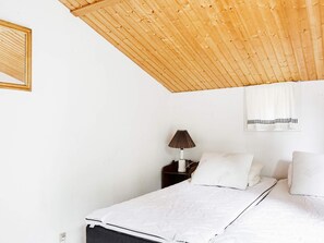 Ein Schlafzimmer mit Holzbalkendecke, einem Bett mit weißer Bettwäsche, einem Nachttisch mit Lampe und einem Fenster mit Vorhang.