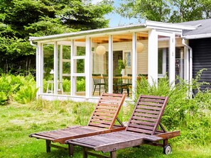 Een overdekt terras met twee houten loungestoelen en een tafel, omgeven door weelderige begroeiing.