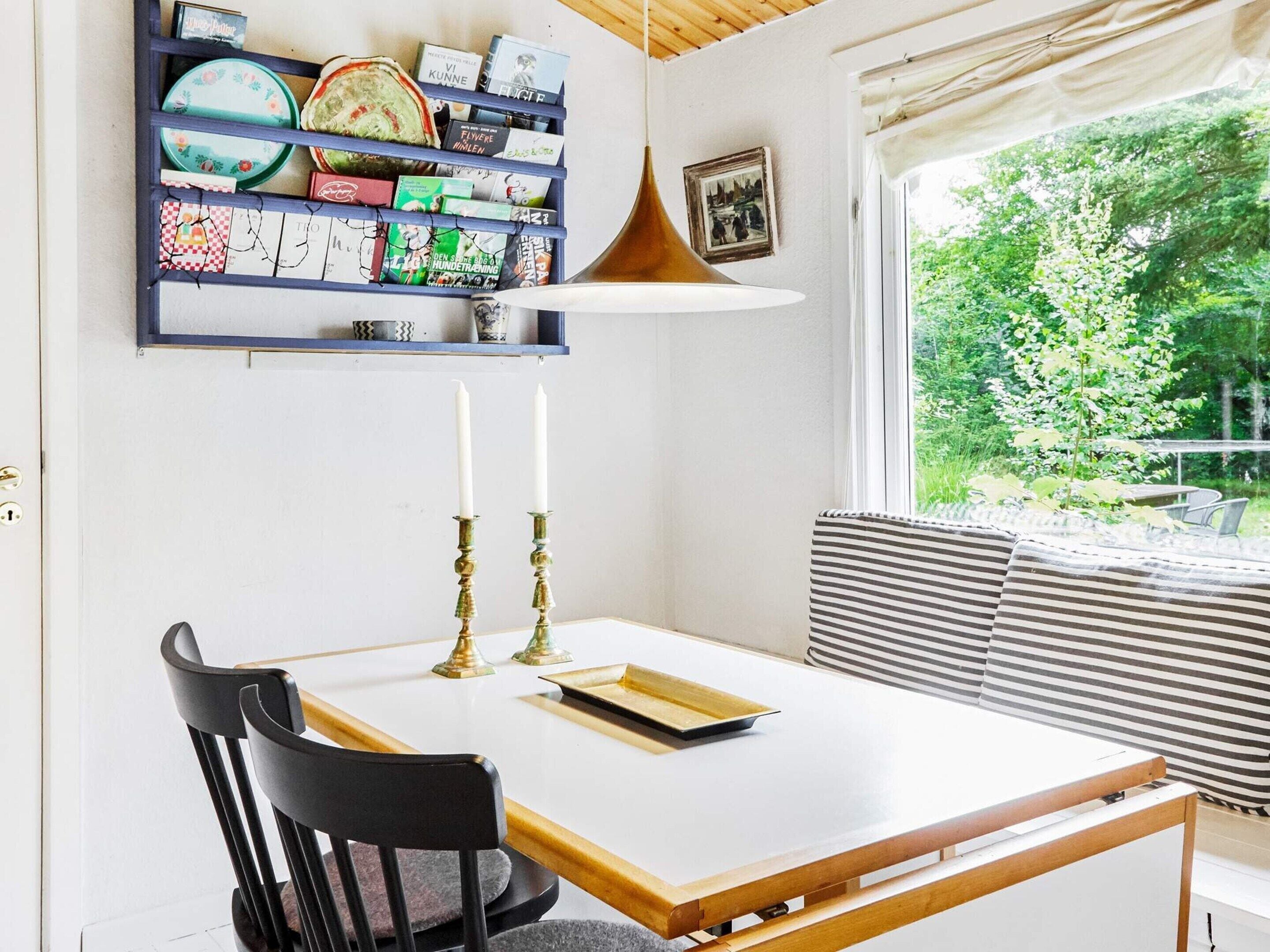 A dining area with a white table, black chairs, a bookshelf with books, and a window with a view of greenery.