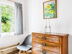 Ein Zimmer mit einem Holzkasten, einem grauen Sessel, einem Fenster mit Vorhängen und einem Sonnenblumenbild.