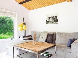 Ein Wohnzimmer mit Holzboden, einem Sofa mit vielen Kissen, einem Couchtisch mit einer Vase und Büchern sowie einem Fenster mit Blick auf Grünfläche.