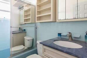 Ein Badezimmer mit Toilette, Waschbecken und Spiegel. Über dem Waschbecken befinden sich Regale und ein Duschbereich mit Glastür.