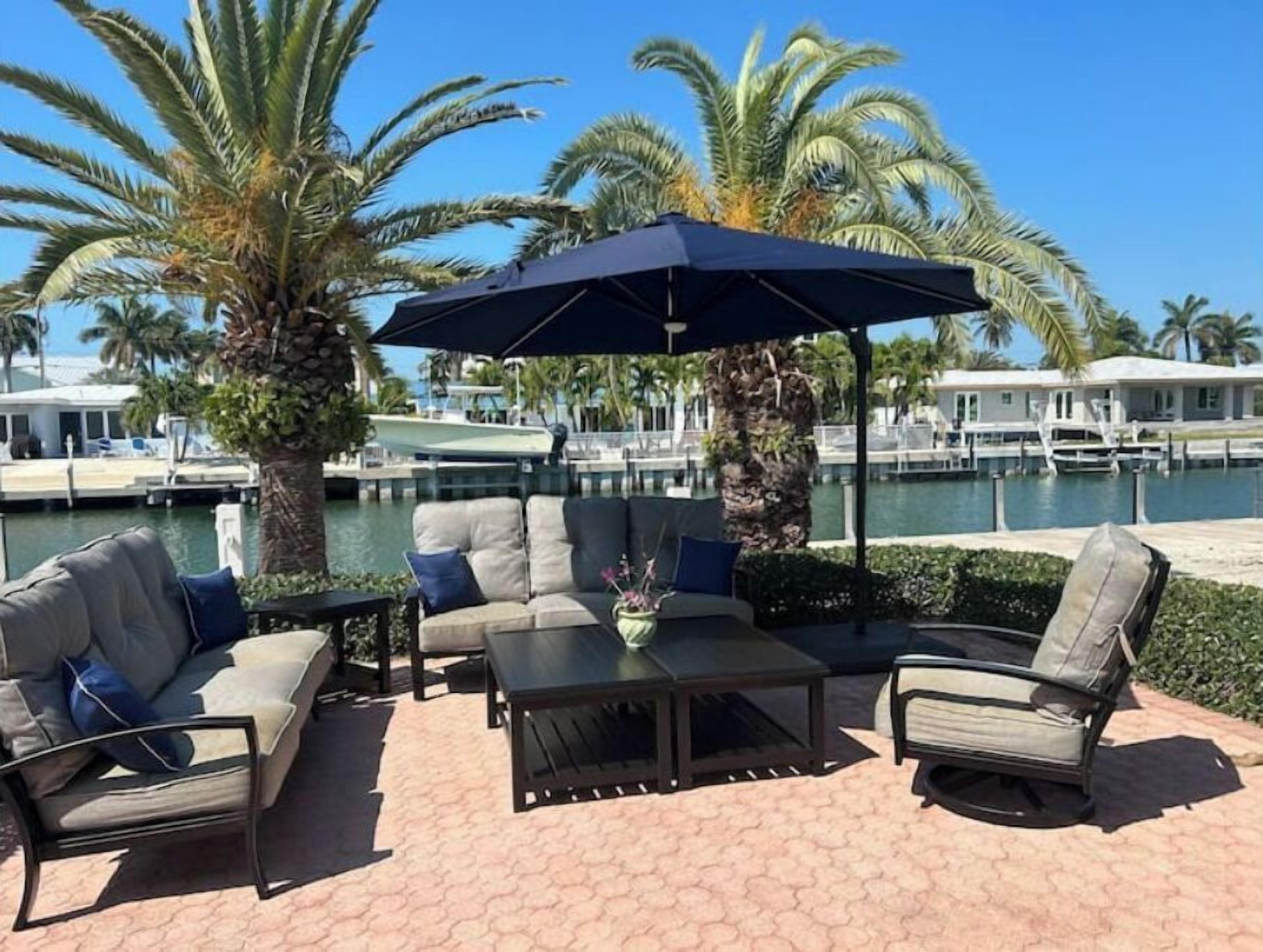 An outdoor seating area with a sofa, chairs, and a table, overlooking a marina with boats and palm trees.
