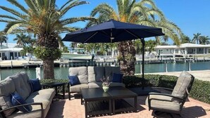 Un espace extérieur aménagé avec un canapé, des fauteuils et une table, offrant une vue sur une marina avec des bateaux et des palmiers.